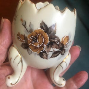 May include: Cream-colored ceramic egg cup with a floral design featuring brown and gold roses. The egg cup has three curved legs and a scalloped rim, with gold trim accents.