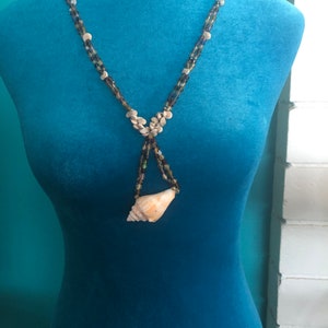 May include: A necklace made of brown, green, and blue beads with a seashell pendant. The necklace is displayed on a blue mannequin.