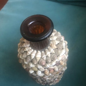 May include: A decorative bottle covered in small seashells. The bottle has a dark brown neck and opening. The seashells are various shades of white, tan, and gray, creating a textured surface. The bottle is likely used for decorative purposes.