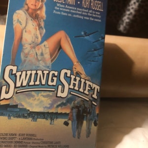 May include: A VHS video cassette cover for the movie "Swing Shift" starring Goldie Hawn and Kurt Russell. The cover features a blue background with a plane flying overhead and a woman in a white dress sitting on a factory floor. The text on the cover reads "Swing Shift" in large white letters.
