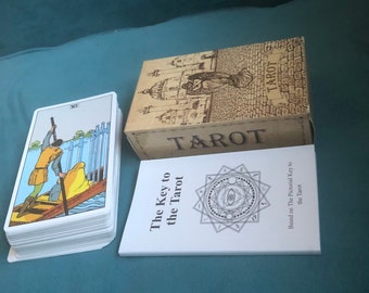 May include: A deck of tarot cards with a white box and a white booklet titled "The Key to the Tarot". The box has a brown background with a black and white illustration of a building and the word "TAROT" in black.