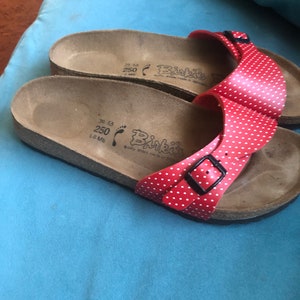 May include: Red polka dot sandals with a black buckle and cork soles. The sandals have the brand name "Binkis" printed on the soles. The size is 250.