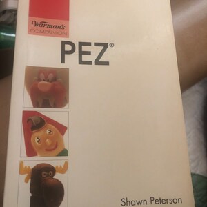 May include: A white book with a red spine and the title "Warman's Companion PEZ®" in black text. The book cover features three cartoon characters: a red monster, a yellow jester, and a brown moose. The author's name, "Shawn Peterson", is printed in black at the bottom of the cover.