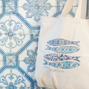 May include: A white canvas tote bag with blue and white patterned fish designs and the text "FARO|PORTUGAL". The bag is sitting on a blue and white tiled surface.