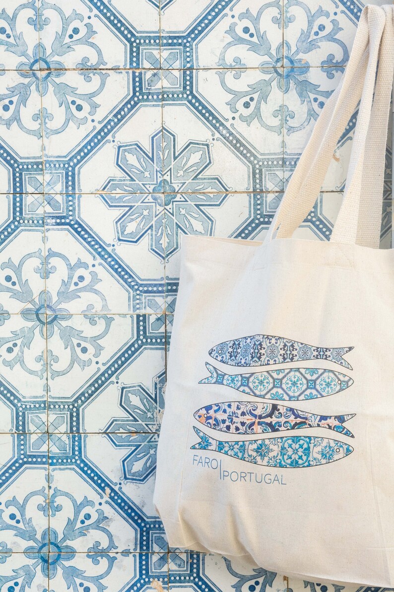 May include: A white canvas tote bag with blue and white patterned fish designs and the text "FARO|PORTUGAL". The bag is sitting on a blue and white tiled surface.