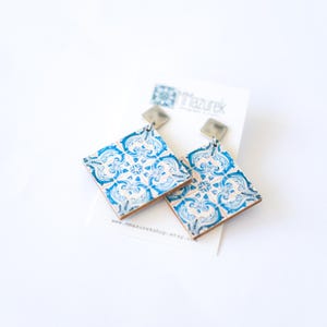 May include: A pair of square earrings with a blue and white tile pattern. The earrings have a silver-colored top and are displayed on a white card with text. The earrings are a unique accessory.