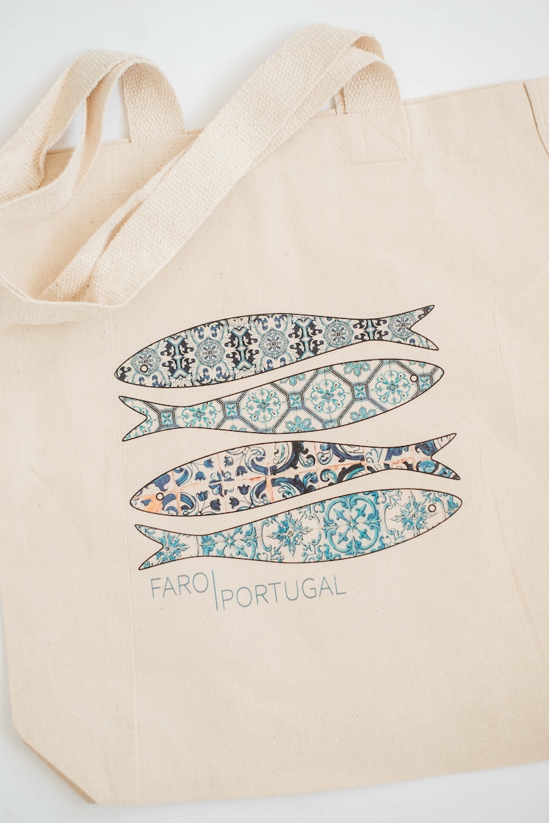 May include: A beige canvas tote bag with blue and white patterned fish and the text "FARO PORTUGAL" printed on the front.
