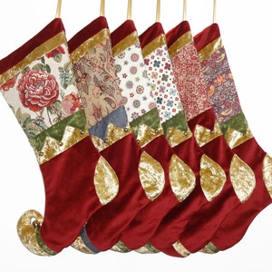 May include: Six Christmas stockings with patterned tops, including floral and paisley designs. The main body of each stocking is a deep red velvet, with gold accents and a gold loop for hanging. The stockings are suspended against a white backdrop.