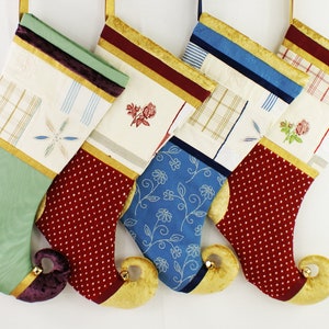 May include: Four decorative Christmas stockings with curled toes and small bells. Each features a cream-coloured cuff with varied patterns, including plaid, floral embroidery, and stripes. The lower sections are made of green, red, and blue fabrics.