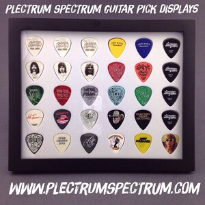 May include: A black framed display case with a white background showcasing a collection of guitar picks. The picks feature various band logos, names, and designs, including "S2000", "John Mayer", "The Best There's Ever Been", "Blando", and "Spike Cassidy DRI".