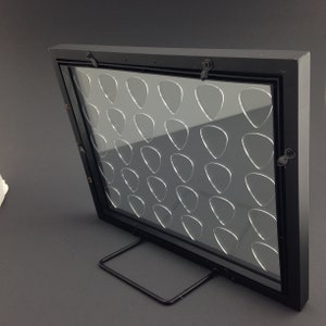 May include: Black guitar pick display frame with a clear acrylic insert and a black metal stand. The frame has 36 slots for guitar picks.