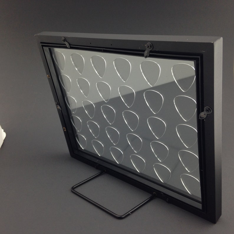 May include: Black guitar pick display frame with a clear acrylic insert and a black metal stand. The frame has 36 slots for guitar picks.