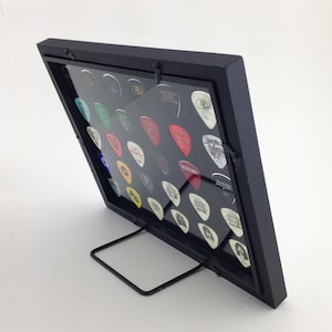 May include: Black guitar pick display frame with a clear acrylic front and a black metal stand. The frame holds a collection of guitar picks with various designs and colors.