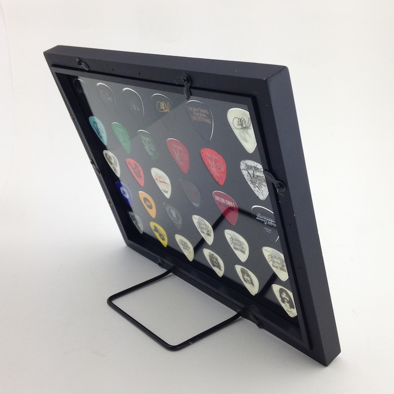 May include: Black guitar pick display frame with a clear acrylic front and a black metal stand. The frame holds a collection of guitar picks with various designs and colors.