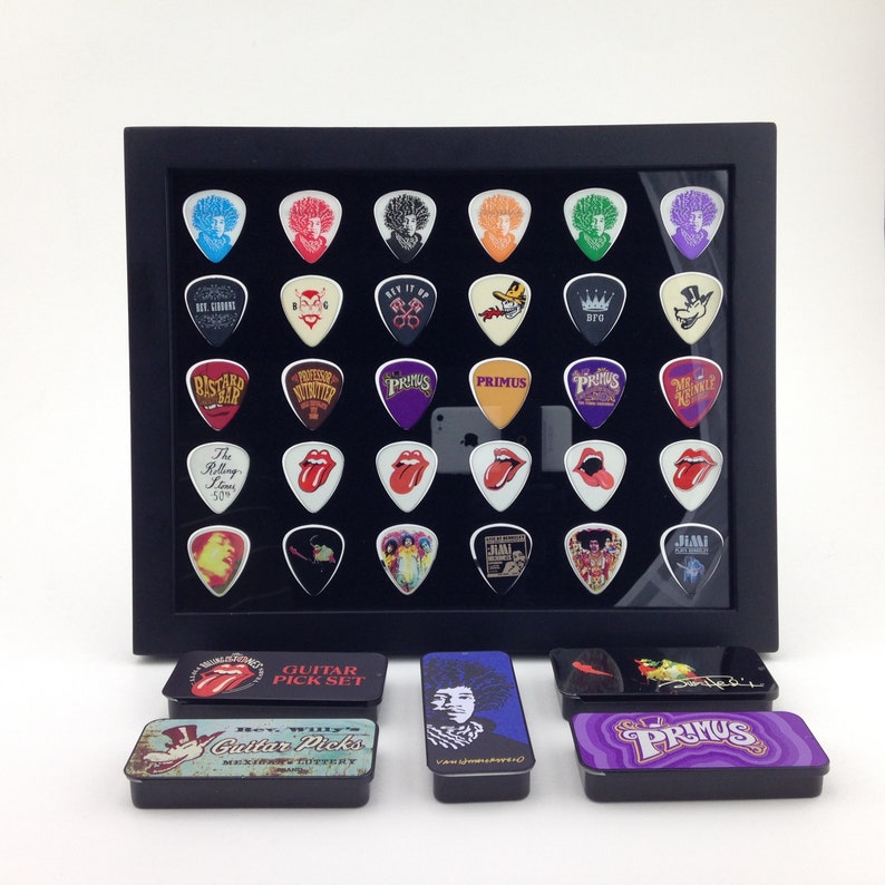 May include: A black frame with 36 guitar picks displayed in a grid pattern. The picks feature various band logos and designs, including Primus, Jimi Hendrix, and The Rolling Stones. There are also three small metal boxes with guitar picks in them. The boxes are labeled "Guitar Pick Set", "Hey Willy's Guitar Picks", and "Primus".