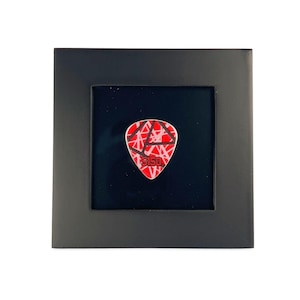 May include: A framed guitar pick with a red and white design and the text "5150". The pick is displayed on a black background within a black frame. The pick has a triangular shape.