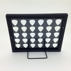 May include: Black framed display stand with a clear acrylic insert and a grid of 36 white guitar picks. The stand has a metal kickstand.