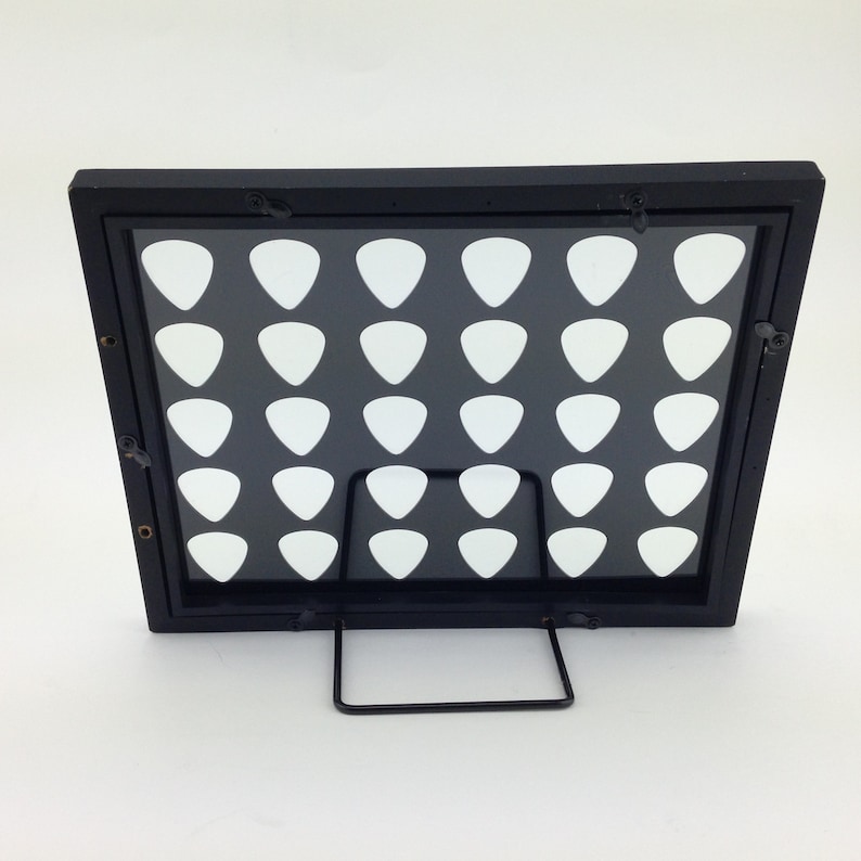 May include: Black framed display stand with a clear acrylic insert and a grid of 36 white guitar picks. The stand has a metal kickstand.