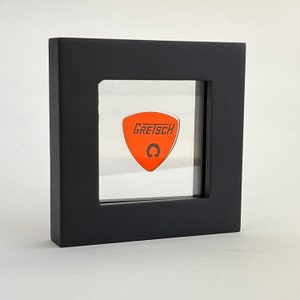 May include: A black display case with a clear front panel showcasing an orange guitar pick with the word "GRETSCH" and a stylized "G" logo.