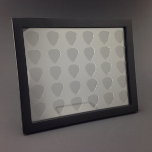 Op de afbeelding: Een zwarte ingelijste vitrine met een transparante glazen voorzijde. De vitrine heeft 25 uitsparingen in de vorm van gitaarplectrums.