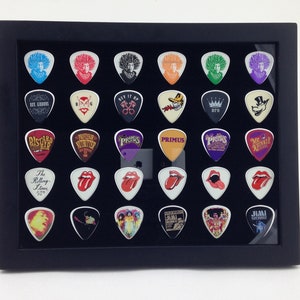 May include: A black framed display case with a black velvet background holds 30 guitar picks. The picks feature various band logos and designs, including The Rolling Stones, Jimi Hendrix, and Primus.