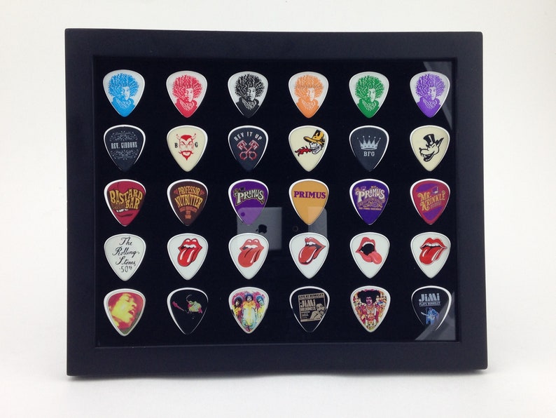 May include: A black framed display case with a black velvet background holds 30 guitar picks. The picks feature various band logos and designs, including The Rolling Stones, Jimi Hendrix, and Primus.