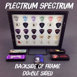May include: A black frame with a clear acrylic front displaying a collection of guitar picks. The picks feature various band logos, including "Primus", "The Rolling Stones", and "KISS". The frame is double-sided and has a black stand.