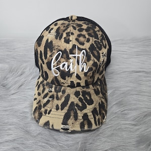 May include: A distressed baseball cap with a leopard print design in shades of brown and black. The word "faith" is embroidered in white script across the front. The back of the cap is black mesh.