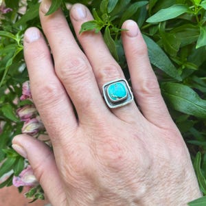May include: A silver ring featuring a square-shaped turquoise stone with dark matrix veining. The stone is set in a smooth silver bezel surrounded by a raised, textured silver frame with a square outline, creating a bold and geometric appearance.