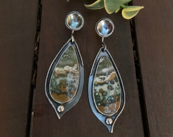 May include: A pair of handcrafted silver earrings with teardrop-shaped, patterned stone insets. The stones feature a mix of green, brown, and white hues. Each earring has a small, round silver accent at the bottom and a larger, clear, round accent at the top.