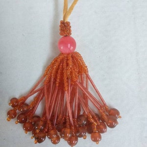 May include: A brown and orange tassel with a pink bead at the top. The tassel is made of small beads and has a loop at the top for hanging.