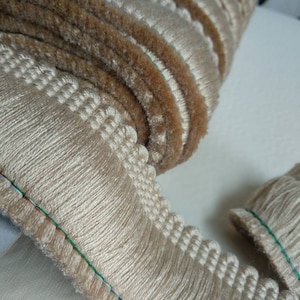 May include: Close-up of a roll of decorative trim. The trim features a light beige fringe with a scalloped edge and a brown, textured rope detail. The trim is likely used for upholstery or crafting projects.