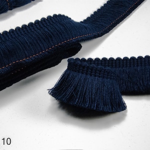 Pode incluir: Close-up de uma guarnição de franjas azul escuro com um detalhe de costura laranja sutil. A guarnição é dobrada ao meio e a franja é visível em ambos os lados. A guarnição é etiquetada como "Col. 10".