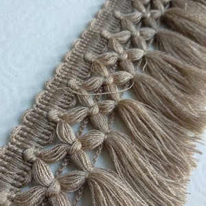 May include: A close-up of a beige tassel trim with a woven pattern. The trim has a fringe of tassels that are about 1 inch long.