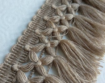 May include: A close-up of a beige tassel trim with a woven pattern. The trim has a fringe of tassels that are about 1 inch long.