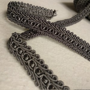 May include: Grey trim with a decorative, braided design. The trim has a repeating pattern of loops and knots.