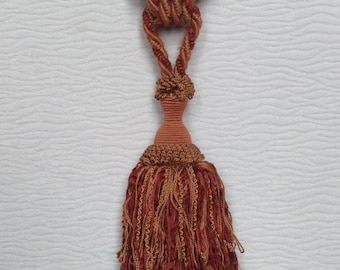 May include: A brown and gold tassel tieback with a fringed bottom. The tieback is made of a woven cord and has a decorative knot at the top.