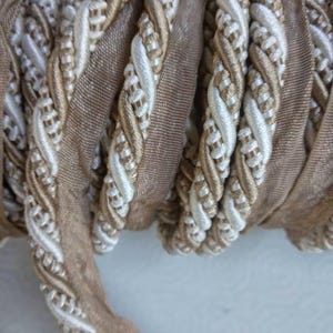 May include: A spool of 10mm or 0.39 inch flanged piping cord featuring a twisted design in ivory and beige. The trim consists of a flat fabric flange attached to a decorative braided cord, showcasing a neutral color palette and a textured finish.