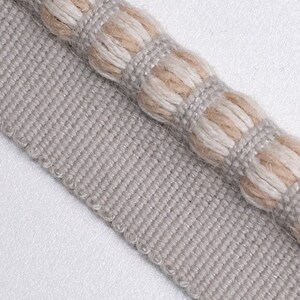 May include: A close-up of a gray and beige trim with a textured, woven design. The trim has a raised, rope-like detail along the edge.