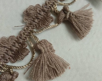 May include: A brown tassel trim with a woven design and a gold cord running through it. The trim has a series of tassels that hang down.