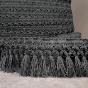 May include: Grey crocheted trim with a fringe. The trim has a textured pattern and is made of a soft, woven fabric.