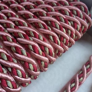 May include: Close-up of a braided trim with a pink and burgundy color scheme. The trim is made of a soft, woven fabric and has a subtle sheen.