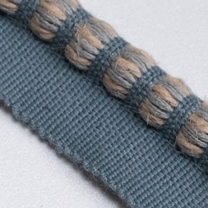 May include: Close-up of a blue and beige fabric trim with a textured, rope-like design. The trim has a woven base with a raised, knotted pattern.