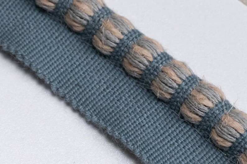 May include: Close-up of a blue and beige fabric trim with a textured, rope-like design. The trim has a woven base with a raised, knotted pattern.