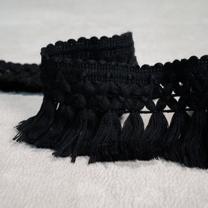 May include: Black tassel trim with a woven design. The trim has a fringe of tassels along the bottom edge.