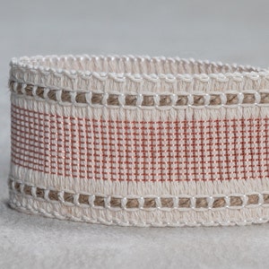 May include: Close-up of a white and brown woven trim with a thin pink stripe. The trim has a textured, woven design.