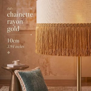 Op de afbeelding: Een goudkleurige vloerlamp met een crèmekleurige lampenkap, afgewerkt met gouden franjes. De tekst "chainette rayon gold" is zichtbaar. De lampenkap heeft een diameter van 10 cm. Een turquoise kussen met een crèmekleurige rand ligt op de vloer.