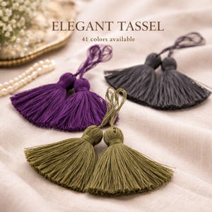 Pode incluir: Close-up de três pares de borlas elegantes em tons de roxo, cinza e verde-oliva. A imagem inclui o texto "ELEGANT TASSEL" e "41 cores disponíveis". As borlas são feitas de um material macio e fluido.
