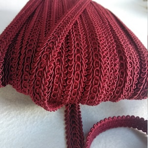 Puede incluir: Borde de trenzado rojo burdeos con un patrón de bucles decorativos. El borde está enrollado y una pequeña sección está dispuesta en plano.