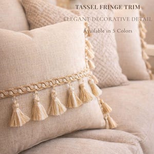 Puede incluir: Cojín decorativo beige con ribete de flecos de borlas. El cojín tiene un elegante detalle decorativo y está disponible en cinco colores. Las borlas son de color beige claro y cuelgan de un adorno decorativo.
